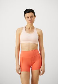 Sujeción deportiva en rosa claro con logo blanco y tirantes anchos, combinada con pantalones cortos de talle alto en naranja brillante que presentan detalles de bolsillo lateral.