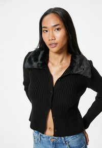 Cardigan nero a coste con un colletto nero in peluche, caratterizzato da un design aderente e chiusure a bottone, abbinato a jeans in denim blu.