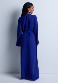 Robe longue et fluide de couleur bleue en tissu transparent, avec des manches longues, une taille ceinturée et un détail en forme de clé à l'arrière.