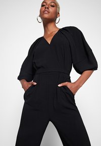 Combinaison noire avec un décolleté en V, des manches bouffantes et une taille élastique. Dispose de poches latérales et d'une texture lisse avec une silhouette cintrée.