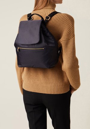 Mochila texturizada azul marino, con cierre de solapa y bolsillo frontal con cremallera. Tiras ajustables y acentos de hardware dorado. Llevada sobre un suéter de canalé.