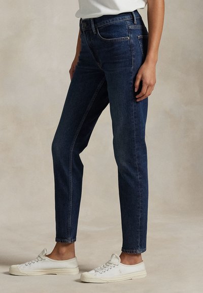 Osoba nosi tmavomodré priliehavé džínsy a biele nízke tenisky, stojaca pred neutrálnym béžovým pozadím.