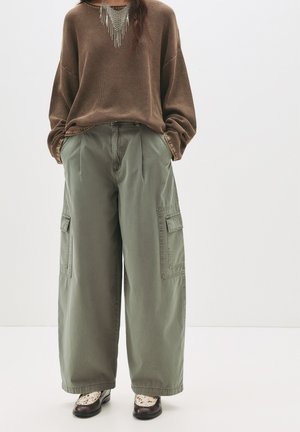 Pull en maille marron à manches longues associé à un pantalon cargo oversize vert olive avec des poches latérales. Les chaussures sont à motifs noir et blanc.