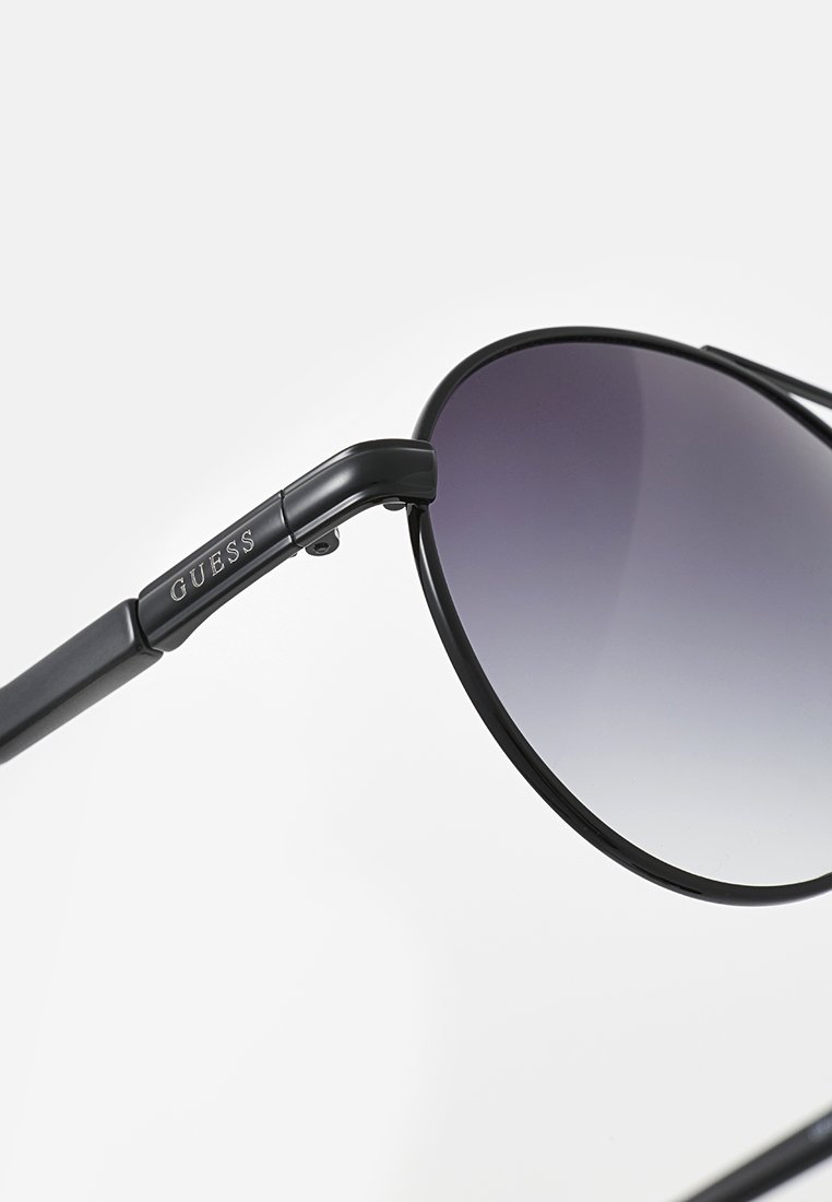 Occhiali da sole neri con lenti sfumate. Montatura in metallo sottile con una finitura lucida e logo "GUESS" sull'asta. Forma delle lenti rotonda.