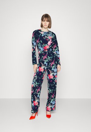 Macacão floral com mangas longas em azul-marinho, apresentando uma mistura de flores rosa, vermelhas e azuis; combinado com sapatos de salto alto vermelhos brilhantes.