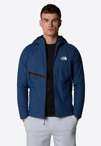 Blå isolerad jacka med huva, framdragkedja och en bröstficka med svart dragkedja. Logotypen visas på vänster bröstområde.