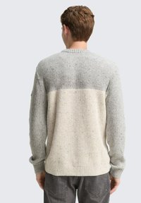 Mann, der einen grau und cremefarbenen, blockfarbigen Strickpullover trägt, mit dem Rücken zur Kamera, kombiniert mit dunkelgrauen Jeans, vor einem schlichten, hellen Hintergrund.