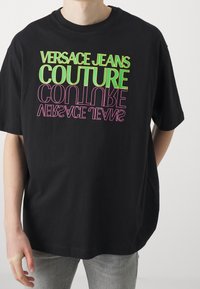 Svart bomullströja med fet flerfärgad text: "VERSACE JEANS COUTURE" i grönt och rosa, med korta ärmar och avslappnad passform.