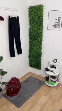 Fekete nadrágok lógnak egy fehér állványról, mellette egy függőleges zöld növényfal áll. Egy piros táska pihen egy szürke szőnyegen egy fehér varrógép és könyvek mellett.