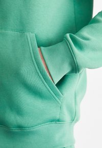 La mano di una persona parzialmente dentro la tasca anteriore di una felpa verde, in evidenza il tessuto e i dettagli della cucitura.