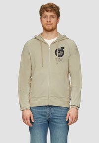 Sweatshirt beige à capuche avec fermeture éclair sur le devant, grandes poches avant et impression graphique du logo "The Big Apple". Texture douce, coupe décontractée.