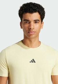 Camiseta amarilla claro de manga corta con cuello redondo y un logo de tres rayas en gris oscuro en el pecho, hecha de tela suave.