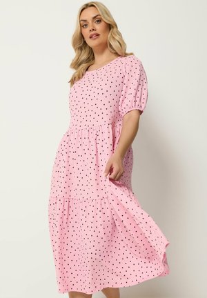 Femme portant une robe midi rose à pois noirs, manches courtes bouffantes et jupe évasée à volants, debout devant un fond uni.