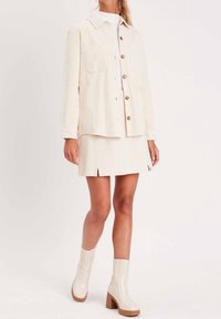 Ljusbeige kordjacka med knappstängning och två fickor, i kombination med en matchande minikjol med en slits fram. Vita ankelstövlar med kraftig klack.