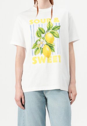 Person, die ein weißes T-Shirt mit einem Zitronenzweig, Blumen und dem Text „Sauer & Süß“ trägt, kombiniert mit hellblauen Jeans.