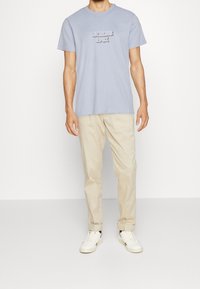 Camiseta de algodón azul claro con el logotipo en relieve "GUESS 1981", combinada con pantalones beige y zapatillas blancas. Diseño sencillo de cuello redondo.