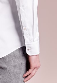 Camisa blanca de manga larga con textura suave, con botones de perla en los puños y un diseño entallado, combinada con pantalones grises con textura.
