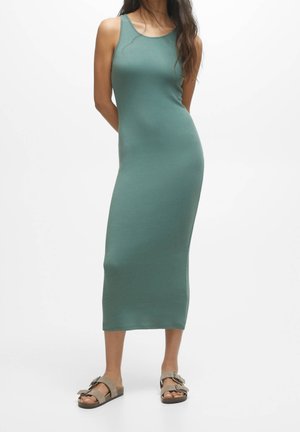 Mujer que lleva un vestido largo sin mangas de color azul verdoso y sandalias beige con hebilla, de pie frente a un fondo blanco liso.