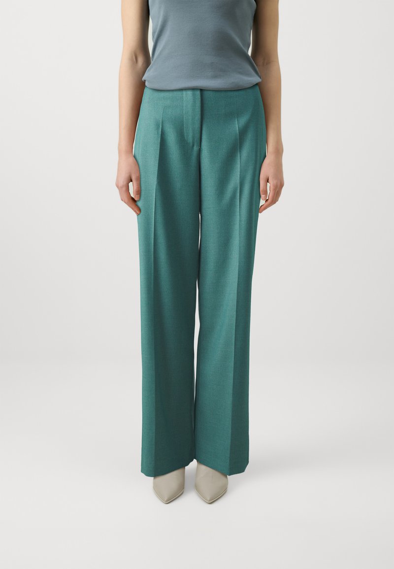 Vidde bukser i teal stoff med tekstur, med frontfolder. Kombinert med en tettsittende lyseblå ermeløs topp og beige sko.