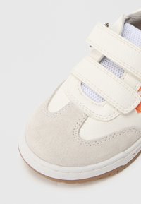 Balti ir pilki sportiniai bateliai iš zomšos ir sintetinės odos. Juose yra lipnūs dirželiai, tinkliniai skydeliai ir guminė pado su smėlio spalvos akcentais.