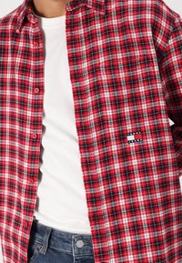 Czerwona i czarna flanelowa koszula w kratę z białymi akcentami, zapinana na guziki, z naszywką z marką na lewej piersi. Noszona na białej koszulce.