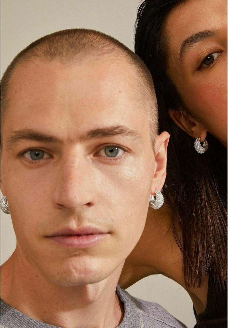 Pendientes de aro plateados con una superficie texturizada, que presentan un diseño brillante. Primer plano de una persona que lleva pendientes, vestida con un suéter gris.