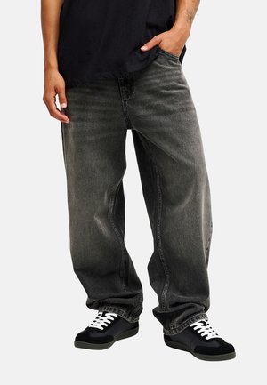 Personne portant un jean gris foncé ample, des baskets noires avec des lacets blancs et une chemise noire, une main dans la poche.