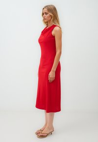 Rode mouwloze jurk met een drapering bij de hals, knielange snit, gladde textuur en slanke pasvorm, gecombineerd met eenvoudige nude sandalen.