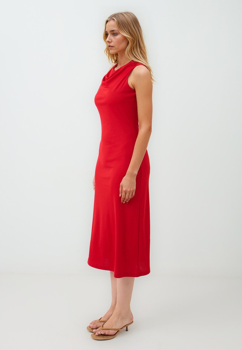 Rode mouwloze jurk met een drapering bij de hals, knielange snit, gladde textuur en slanke pasvorm, gecombineerd met eenvoudige nude sandalen.