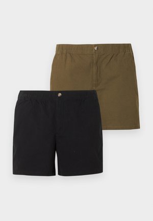 Homme portant un t-shirt marron, un short vert olive, une casquette assortie, des chaussettes grises et des chaussures marron sans lacets, debout sur un fond blanc uni.