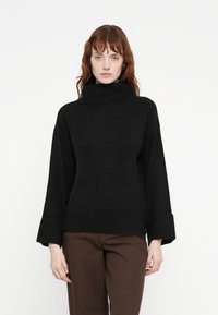 Pull à col roulé surdimensionné noir avec une texture douce et des poignets larges. Assorti à un pantalon droit marron, sur fond blanc.