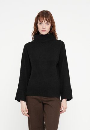 Must have valge suuruse poolest turtleneck särk pehme tekstuuriga ja laiad mansetid. Kombineeritud pruunide sirgete pükstega valge taustaga.