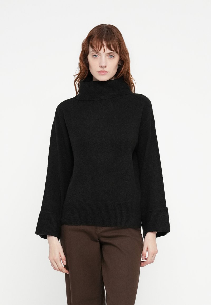 Pull à col roulé surdimensionné noir avec une texture douce et des poignets larges. Assorti à un pantalon droit marron, sur fond blanc.