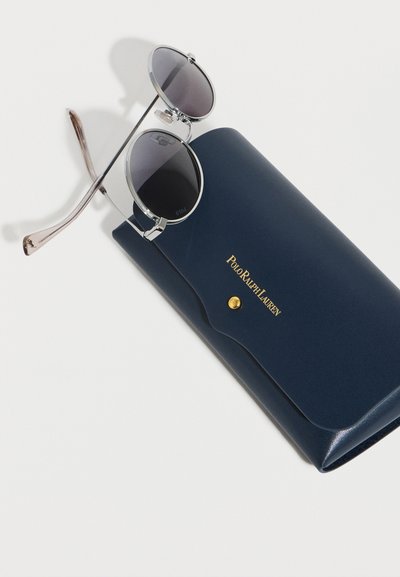 Στρογγυλά γυαλιά ηλίου με ασημένια περιτύλιξη και σκουρόχρωμους φακούς, τοποθετημένα πάνω σε θήκη Polo Ralph Lauren σε ναυτικό μπλε χρώμα με επίχρυσο κουμπί κλεισίματος.