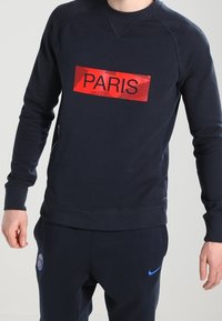 Felpa blu navy con testo rosso "PARIS" e sfondo a righe sul davanti, con polsini e orlo a coste, abbinata a pantaloni della tuta scuri.