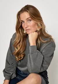 Sudadera gris con detalles de mangas gathered, textura suave y corte relajado, combinada con grandes pendientes metálicos llamativos.