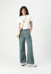 Lys grønne vidde bukser, hvit t-skjorte med grafisk tekst, og beige crossover-veske. Myk tekstur og avslappet passform, parret med lyse sneakers.