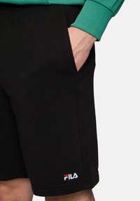 Pantalones cortos de algodón negro con bolsillos laterales, presentando un pequeño logotipo bordado de FILA en rojo y blanco en el dobladillo. Textura suave, diseño casual.