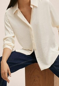 Chemise boutonnée couleur crème avec une coupe décontractée, manches longues retroussées, fabriquée dans un tissu doux et léger. Elle présente un col classique et peu de boutons.