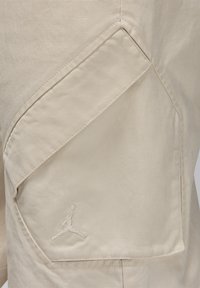 Beige stof med en tekstureret overflade, der har en stor diagonal lomme og et broderet Jumpman-logo i en matchende nuance.