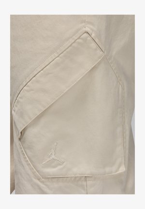 Tissu beige avec une surface texturée, comportant une grande poche diagonale et un logo Jumpman cousu dans une teinte assortie.