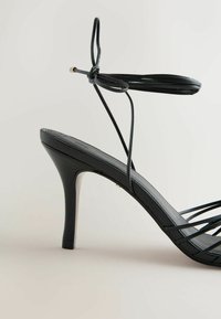 Sandale noire à talon haut avec fines lanières autour de la cheville nouées en nœud, talon aiguille fin et multiples lanières devant sur un fond neutre.