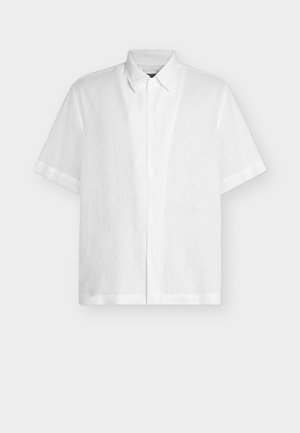 Camisa blanca de manga corta con botones y cuello clásico, mostrada extendida sobre un fondo claro y liso.