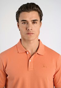 Polo a maniche corte in arancione corallo, realizzato in tessuto morbido, presenta un colletto classico, patta con bottoni e logo ricamato sul petto.