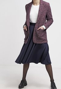 Fialový texturovaný blazer přes bílý pletený svetr, kombinovaný s námořnickou áčkovou sukní a černými botami, s bočními kapsami a dlouhými rukávy.