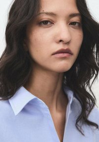 Camicia azzurra con una texture liscia, dotata di bottoni e un colletto strutturato. Il modello ha i capelli ondulati e un trucco naturale.