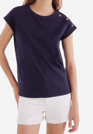 Kvinde iført en mørkeblå kortærmet top med tre knapper på skulderen og hvide shorts med opsmøgede kanter.