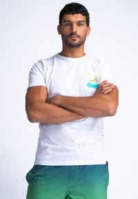 Hvit bomull t-skjorte med et fargerikt grafisk trykk av bølger og skyer på venstre bryst. Modellen har på seg grønne gradient-shorts.