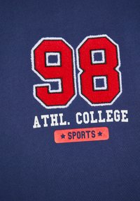 T-shirt in cotone blu navy con numeri rossi in rilievo "98" bordati di bianco e testo bianco "ATHL. COLLEGE SPORTS" sottostante.