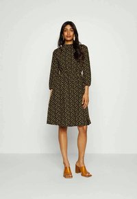 Vestido preto com bolinhas amarelas, decote alto, mangas longas bufantes e cintura ajustada. Usado com sandálias de salto alto em tom de caramelo.